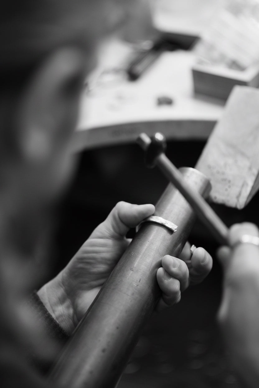 Black and white close-up of a person's hands holding a metal tube while working at a bench with tools in the blurred background.